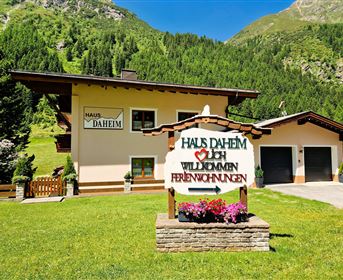 A cozy holiday home in the mountains with a green lawn. A welcome sign in the foreground says "Haus Daheim".