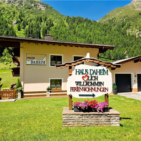 A cozy holiday home in the mountains with a green lawn. A welcome sign in the foreground says "Haus Daheim".