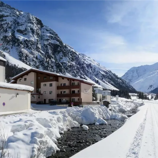 A quiet winter landscape with snow-covered mountains. Nearby, a clear stream flows beside a cozy building.