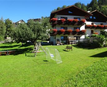 Ein malerisches Haus mit blühenden Balkonen inmitten einer grünen Wiese. Im Vordergrund stehen eine Schaukel und eine Holzbank.