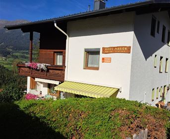 Ein charmantes Haus in den Bergen mit einem blühenden Balkon und einem gelben Sonnenschirm. Die umliegende Landschaft ist grün und malerisch.