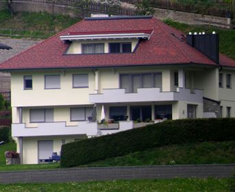 Ein modernes, zweigeschossiges Haus mit rotem Dach und weißer Fassade. Es ist von einem gepflegten Garten umgeben.