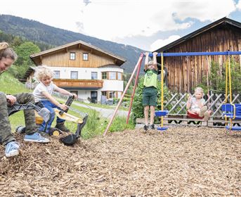 Ein Spielplatz mit Kindern, die draußen spielen. Eine Person sitzt auf einem Spielzeug-Bagger, während andere Kinder schaukeln und sich amüsieren.