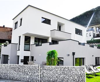 Ein modernes, zweistöckiges Haus mit einer klaren, minimalistischen Fassade. Im Vordergrund sind ein Garten und Liegestühle zu sehen.