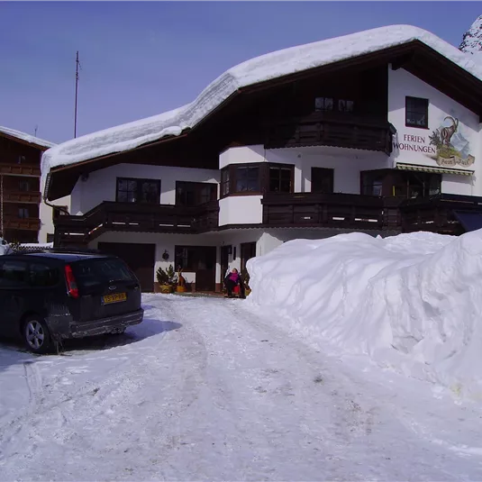 A charming building in the mountains, surrounded by lots of snow. In the foreground, a car is parked while guests are entering the accommodation.