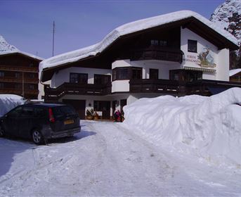 A charming building in the mountains, surrounded by lots of snow. In the foreground, a car is parked while guests are entering the accommodation.