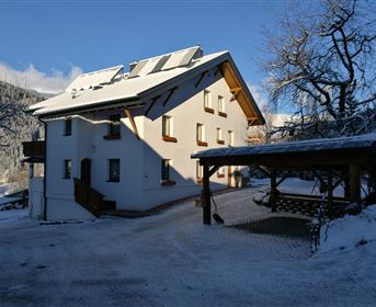 Ein gemütliches Haus im Schnee mit einer Holzüberdachung. Im Hintergrund sind schneebedeckte Berge und blauer Himmel zu sehen.