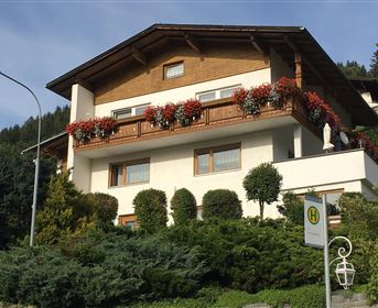 Ein attraktives Haus mit einem Holz-Balkon, der mit bunten Blumen dekoriert ist. Umgeben von grünen Pflanzen und Bäumen.