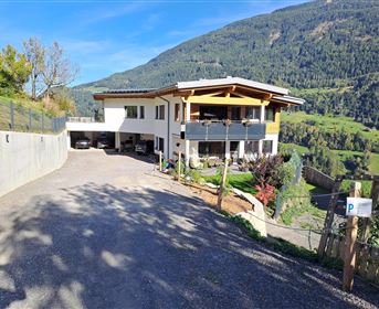 A modern house on a hill with green meadows and mountains in the background. The driveway is paved and surrounded by trees.