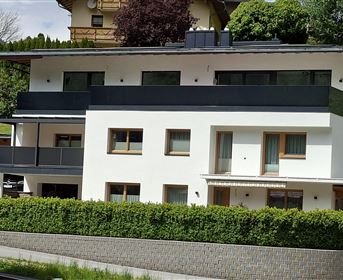 Ein modernes, mehrstöckiges Gebäude mit weißen Wänden und einem schwarzen Balkon. Vor dem Haus befindet sich eine grüne Hecke und Rasenflächen.