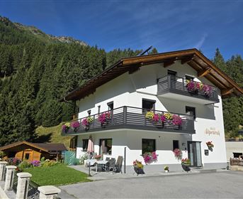 A beautiful, modern house with flower balconies, surrounded by mountains. The sky is clear and blue.