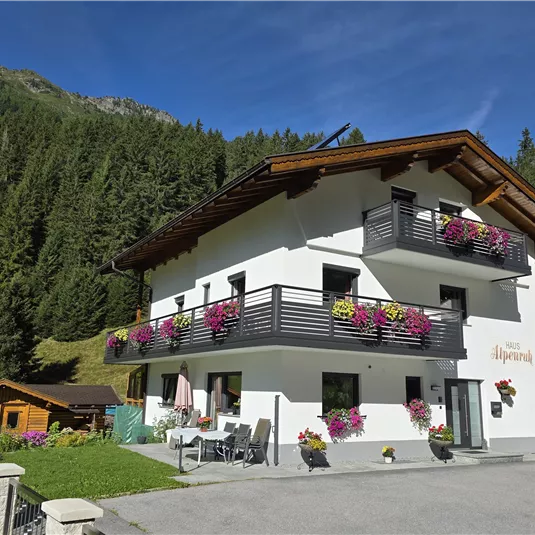 A beautiful, modern house with flower balconies, surrounded by mountains. The sky is clear and blue.