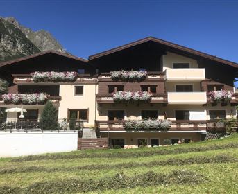 Ein charmantes Gebäude in den Bergen mit bunten Blumen auf den Balkonen. Die Umgebung ist grün und sonnig, umgeben von majestätischen Bergen.