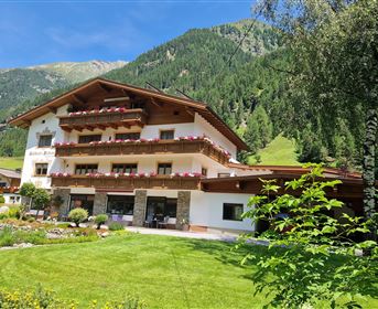 Ein gemütliches Alpenhaus umgeben von grünen Wiesen und Bergen. Der Außenbereich ist mit bunten Blumen und einem gepflegten Garten gestaltet.