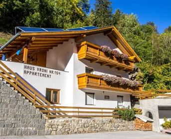 A modern building with wooden cladding and a balcony, surrounded by trees. The house has an inviting entrance area and a well-maintained outdoor space.
