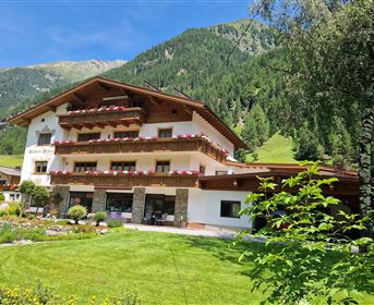 Ein gemütliches Alpenhaus umgeben von grünen Wiesen und Bergen. Der Außenbereich ist mit bunten Blumen und einem gepflegten Garten gestaltet.