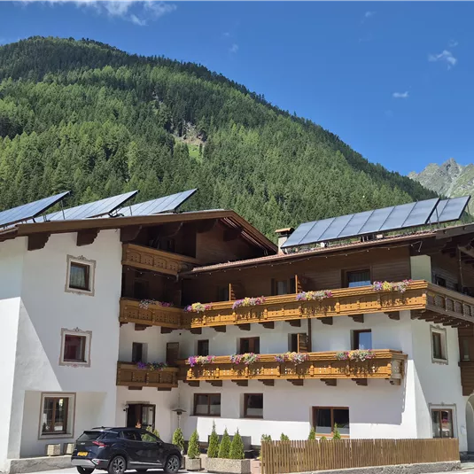 A cozy building in the mountains with balconies and solar panels. The surroundings are filled with green forests and mountains.