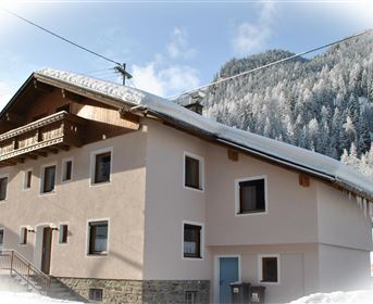 A cozy house in the snow with a snow-covered roof. Forested mountains are visible in the background.