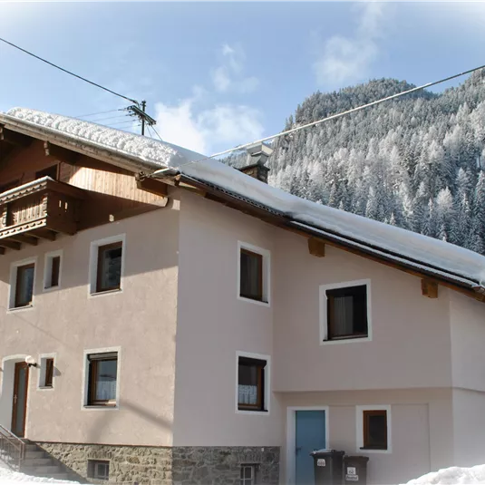 A cozy house in the snow with a snow-covered roof. Forested mountains are visible in the background.