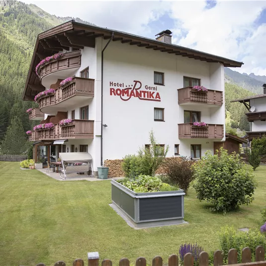 A charming hotel with a garden, surrounded by mountains. Balconies adorned with flowers decorate the facade of the building.