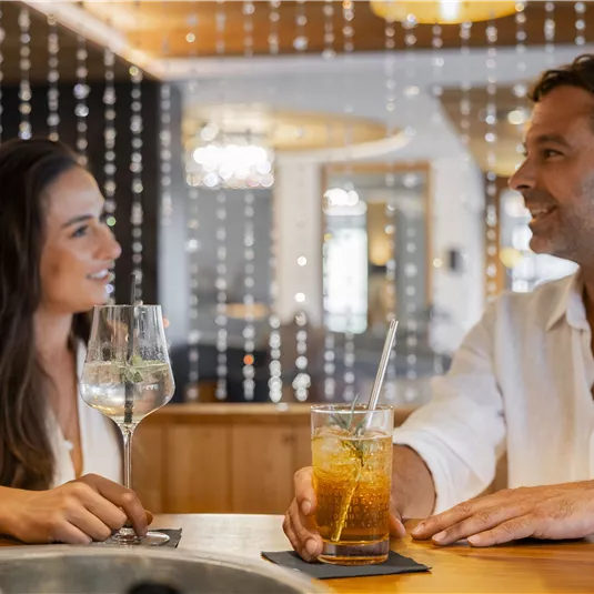 Ein Paar sitzt an der Bar und unterhält sich lächelnd. Auf dem Tisch stehen Getränke, darunter ein Glas mit einem bunten Cocktail.