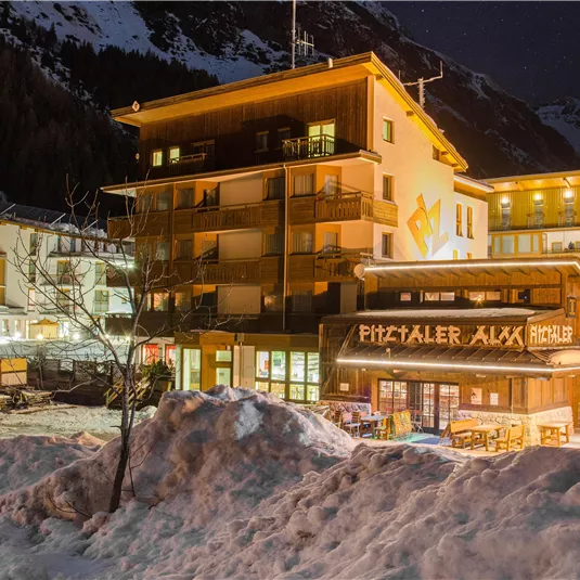 Ein winterliches Dorf mit schneebedeckten Straßen und beleuchteten Gebäuden. Im Vordergrund ist das Restaurant "Pitzaler Axa" sichtbar.