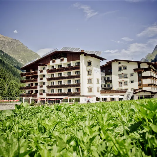 Ein großes Hotel inmitten einer grünen Landschaft und umgeben von Bergen. Der Himmel ist klar und die Atmosphäre ist ruhig und einladend.