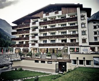Ein elegantes Hotelgebäude in den Alpen mit mehreren Balkonen und schönen Holzverzierungen. Im Vordergrund befinden sich gepflegte Grünflächen und eine entspannte Atmosphäre.