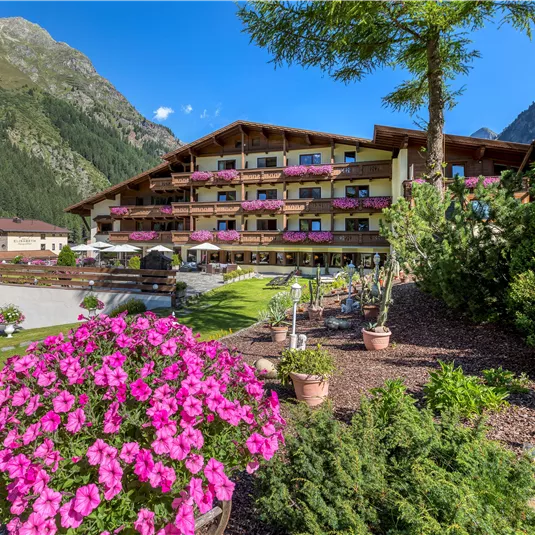 Ein Hotelgebäude umgeben von grünen Bergen und bunten Blumen. Auf den Balkonen blühen pinke Pflanzen und die Umgebung ist einladend.