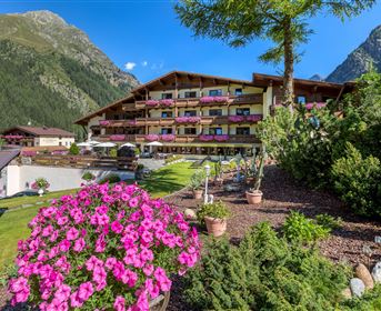 Ein Hotelgebäude umgeben von grünen Bergen und bunten Blumen. Auf den Balkonen blühen pinke Pflanzen und die Umgebung ist einladend.