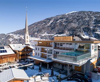 Ein modernes Hotel mit einem Glas-Pool in einer verschneiten Berglandschaft. Im Hintergrund ist eine Kirche mit einem hohen Turm und beeindruckende Berge zu sehen.