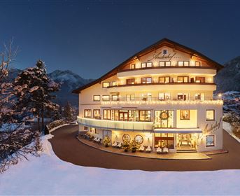 Ein schönes Hotel im Winter mit schneebedeckten Außenterrassen und warmem Licht aus den Fenstern. Die umliegende Berglandschaft ergänzt die einladende Atmosphäre.