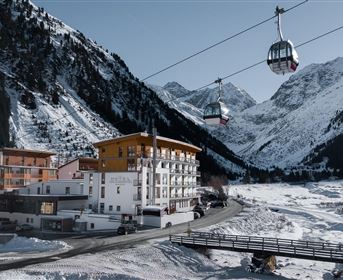Eine schneebedeckte Berglandschaft mit einem modernen Hotel und einer Seilbahn. Die Umgebung ist ruhig und malerisch, umgeben von hohen Bergen.