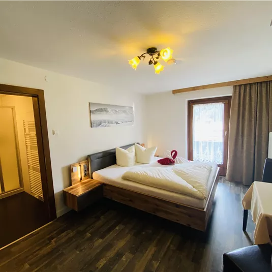 A cozy bedroom with a double bed, wooden floor, and a large window. In the background, there is an en-suite bathroom with a shower.