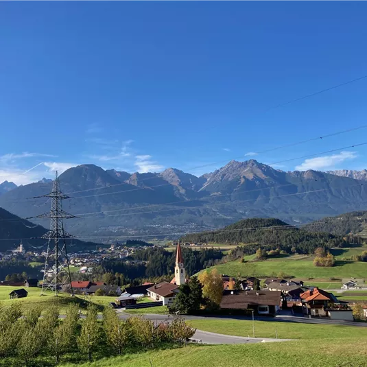 A picturesque landscape with mountains in the background and a small village in the foreground. The sun is shining and the sky is clear and blue.