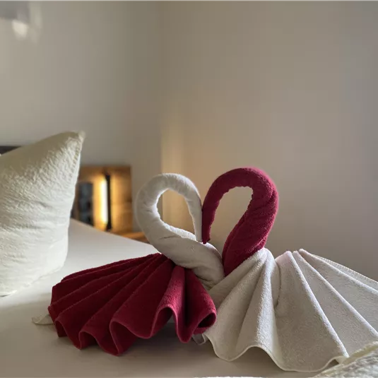 An artistic towel folding that represents two swans. They are designed in red and white and are placed on a bed with pillows in the background.