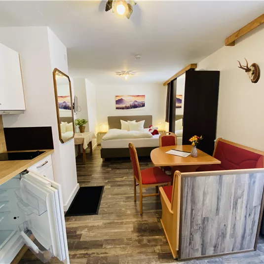 A modern apartment with a cozy dining area and an open kitchen. In the background, a bedroom is visible with a large bed and a beautiful view.