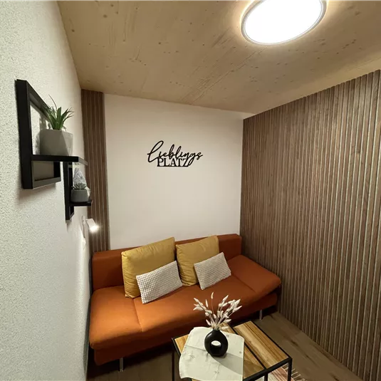 A cozy room with an orange sofa and a small table. Plants and a decorative font hang on the wall.