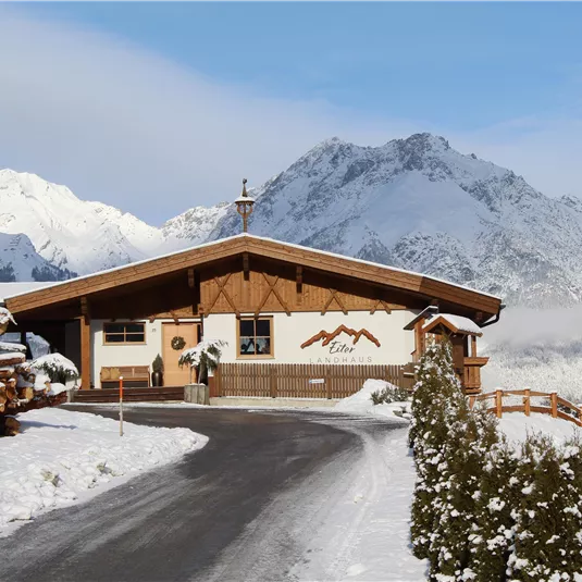 A cozy wooden house in a snowy mountain landscape. Tall mountains and a clear sky can be seen in the background.