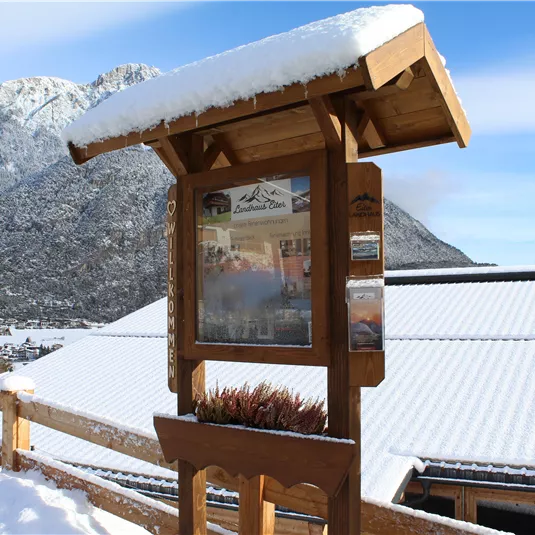 A wooden overview board stands in the snow with a mountain view. The surroundings are wintry and covered in snow.