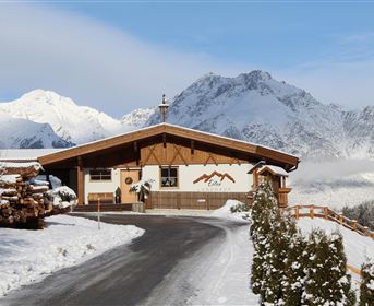 A cozy wooden house in a snowy mountain landscape. Tall mountains and a clear sky can be seen in the background.