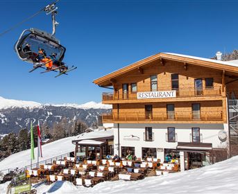 A ski lift that goes over a restaurant in the mountains. In the foreground, there are tables in the snow, and in the background, snow-covered mountains can be seen.