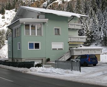 Ein mehrstöckiges, grünes Gebäude in einer schneebedeckten Landschaft. Im Hintergrund sieht man verschneite Bäume und eine ruhige Straße.