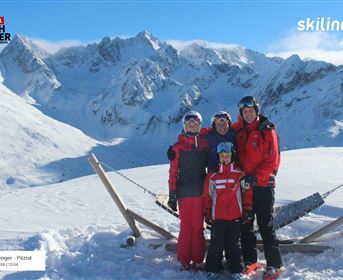 A family stands in the snow in front of impressive mountains. Everyone is in ski clothes and enjoying the sunny day.