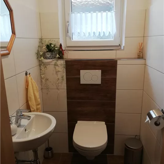 Ein modernes Badezimmer mit einer weißen Toilette und einem kleinen Waschbecken. An der Wand gibt es Dekorationen und ein Fenster sorgt für natürliches Licht.