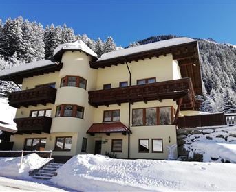 Ein schönes, dreistöckiges Haus im Schnee mit traditioneller Architektur. Im Hintergrund sind schneebedeckte Bäume und ein blauer Himmel zu sehen.