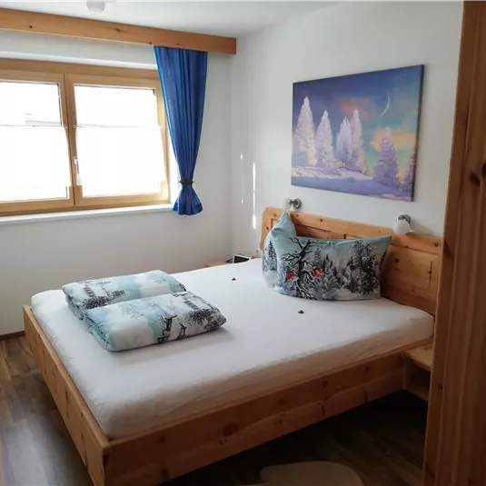 Ein gemütliches Schlafzimmer mit einem Holzbett und bunten Kissen. Große Fenster mit blauen Vorhängen lassen viel Licht herein.