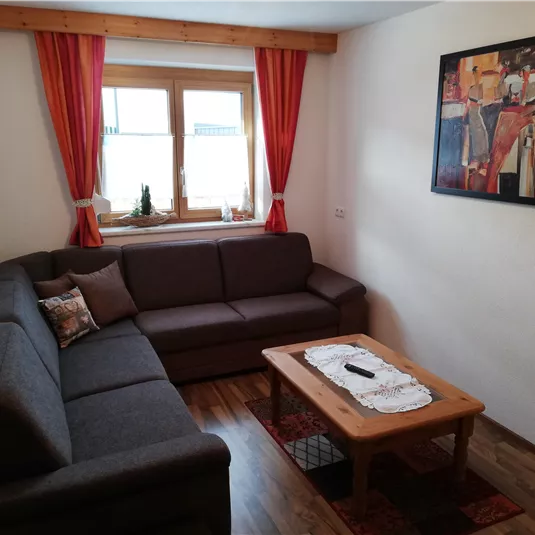Ein gemütliches Wohnzimmer mit einem grauen Sofa und einem Holztisch. Große Fenster lassen viel Licht herein und bunte Vorhänge verleihen dem Raum eine einladende Atmosphäre.