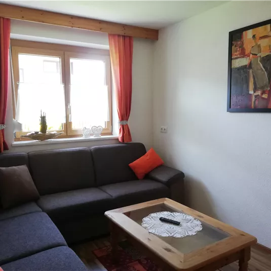 A cozy living room with a gray sofa and orange cushions. A colorful painting hangs on the wall and a wooden table stands in the middle.