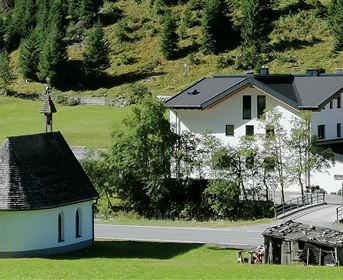 Eine malerische Landschaft mit einer kleinen Kapelle und einem modernen Gebäude. Umgeben von Bäumen und sanften Hügeln.